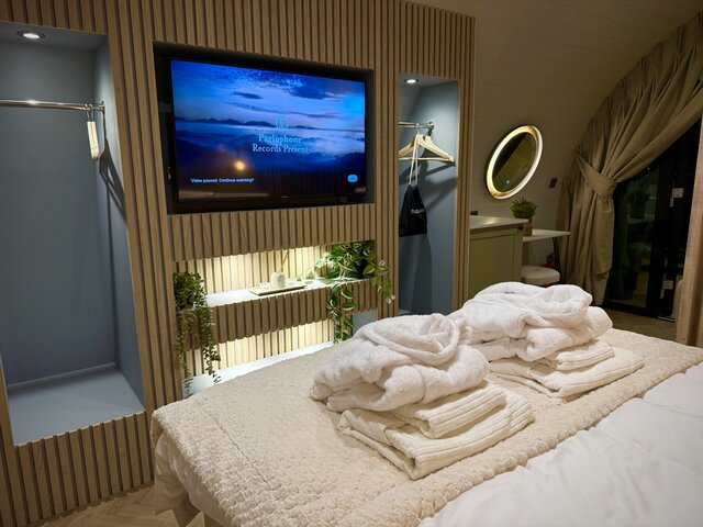 “The Horizon bedroom at The Secret Garden Glamping showing the foot of the bed with neatly arranged towels, blankets, and robes. A full wall-mounted flat-screen TV is visible.