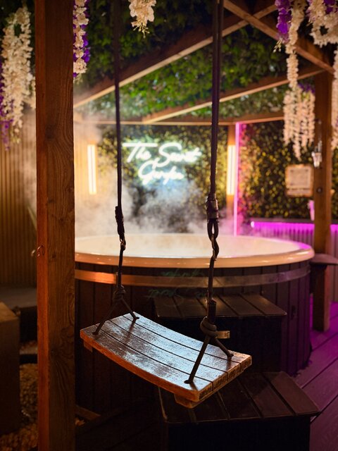 “Luxury outdoor swing with a brown cushioned seat, set in front of a steaming hot tub, creating an inviting and relaxing space ready for guests to unwind in comfort.”