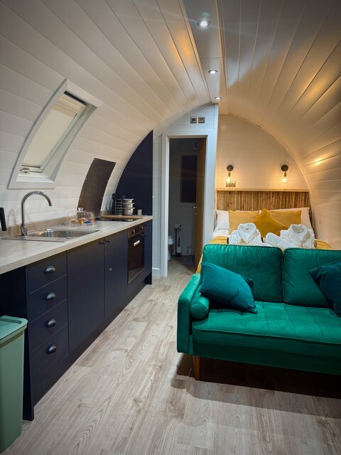 "Interior of a luxury glamping pod at The Secret Garden Glamping, featuring a compact kitchen with black cabinets and a sink on the left, a green velvet sofa in the center.