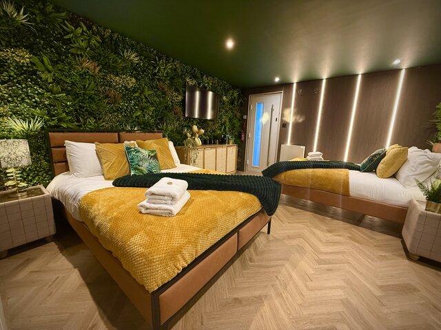 Luxury bedroom with two double beds with yellow and green blankets, beige chevron flooring, bedside tables, jungle-leaf feature wall, and modern brown panel accents at The Secret Garden Glamping.