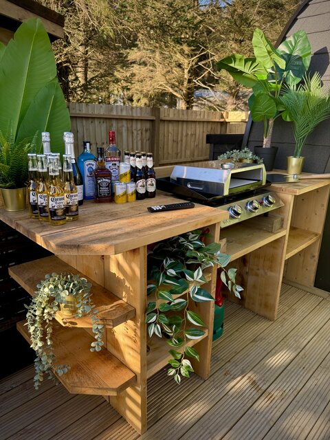“Outdoor bar area at The Cove with a premium hot plate griddle and pizza oven attachment, surrounded by hanging greenery and rustic décor. A selection of luxury spirits and cocktail ingredients.