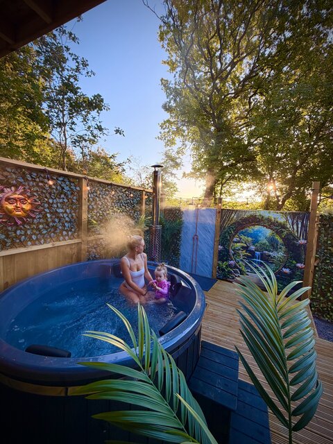 A tender moment unfolds as a mother and daughter relax together in a modern log-burner hot tub, the water glowing a striking bright blue around them. 
