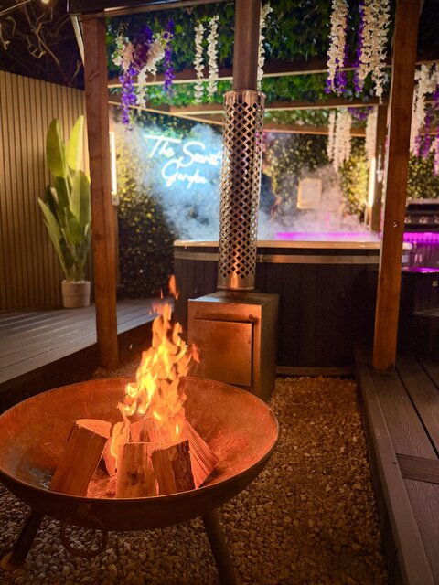 “Roaring outdoor log-burning fire beside a luxury hot tub, perfectly complementing each other to create a warm, indulgent, and inviting outdoor experience.”