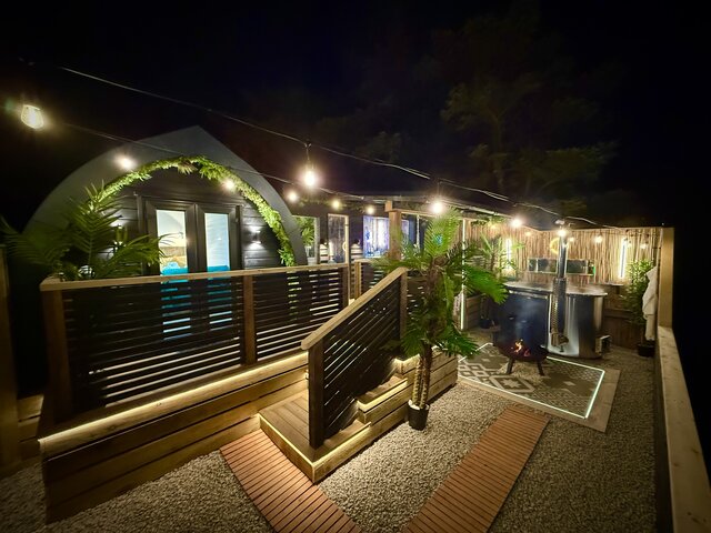 “Nighttime view of the Pavilion pod at The Secret Garden Glamping, featuring a luxury log-burning hot tub in the corner. The pod is illuminated with warm hanging lights, surrounded by lush greenery.