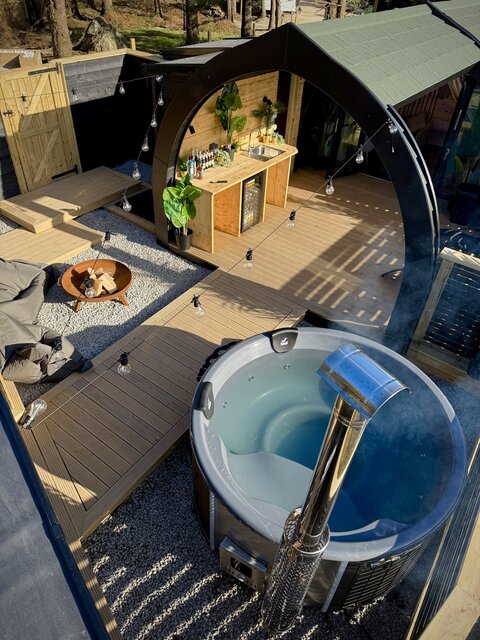 “High drone view of the Horizon pod at The Secret Garden Glamping, showcasing the hot tub with crystal-clear blue water, a fire pit surrounded by pebbles.