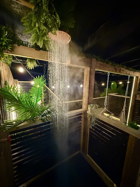 “The Pavilion outdoor shower at The Secret Garden Glamping, with water cascading gracefully and steam rising into the night. Lush greenery hangs from above.
