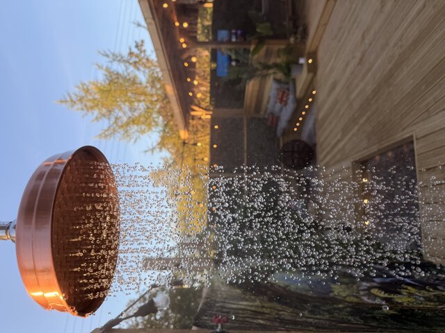 A lavish outdoor shower cascades in a fine, shimmering stream, the droplets catching the warm glow of sunset as they fall. Behind the flowing water, the golden-brown decking stretches out.