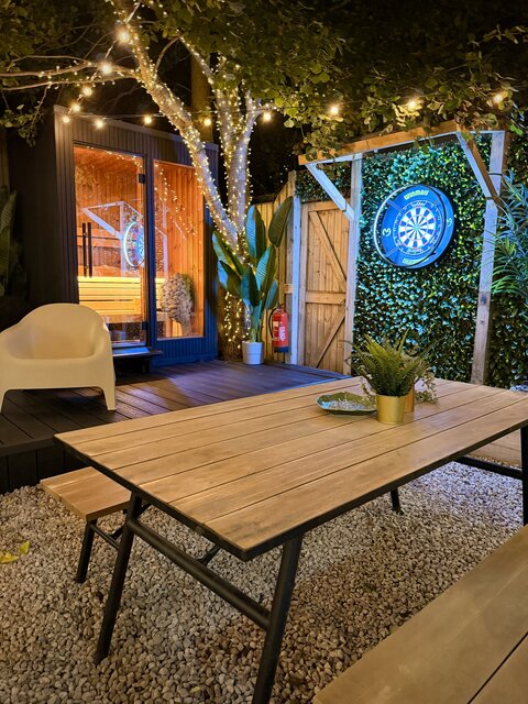 Outdoor area featuring a wooden picnic bench, a private sauna hut and an illuminated dartboard, all set within a cosy, plant-filled setting at The Lodge, Secret Garden Glamping.