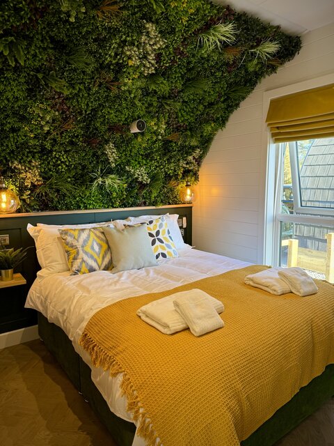 “The Cove bedroom at The Secret Garden Glamping featuring a plush double bed with neatly arranged towels, set against a lush green plant-covered feature wall.