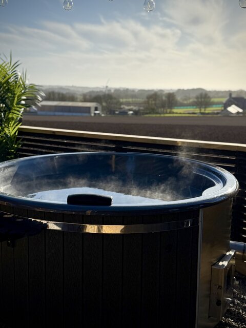 “Outdoor hot tub at The Horizon, The Secret Garden Glamping, overlooking open fields under a blue sky with white clouds. Steam rises gently from the tub, highlighting its luxurious appeal.