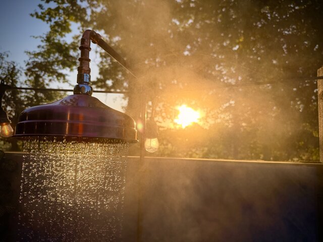 “Luxury outdoor shower at The Secret Garden Glamping with water flowing and steam rising, set against a backdrop of lush trees. The golden hues of the sunset shine through the branches.