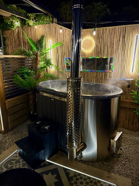 “The Pavilion outdoor log-burning hot tub at The Secret Garden Glamping, with steam rising gracefully into the night and a palm-like plant in the background, creating a serene scene.