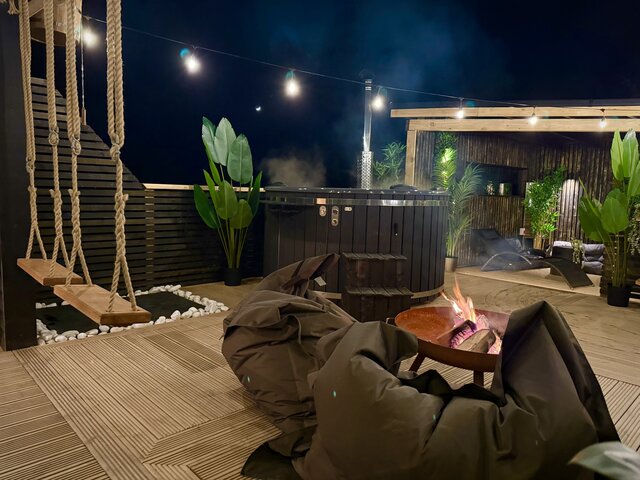 “The Cove outdoor area at The Secret Garden Glamping at night, showcasing cozy striped swinging chairs, a glowing firepit surrounded by plush bag-style seating, and the sleek black hot tub nearby. 