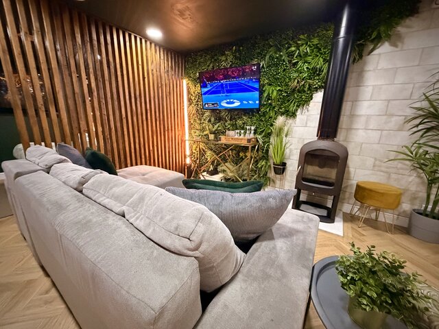 The Lodge living area with beige chevron flooring, a half jungle-leaf and half tiled feature wall, a smart TV, warm stick lighting, a cosy grey corner sofa and a log-burner fire 