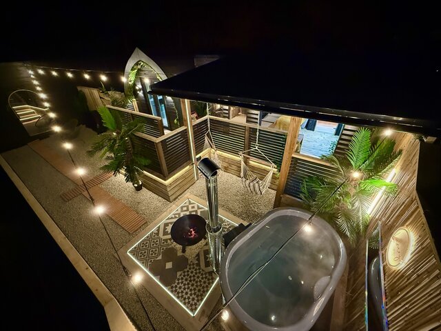 “Drone-style overhead view of The Pavilion outdoor area at The Secret Garden Glamping, featuring a log-burning luxury hot tub filled with bright blue water glowing in the dark. 