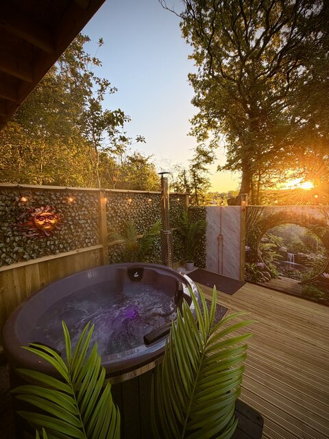 “Luxury log burner hot tub at The Secret Garden Glamping with vibrant blue water, flanked by two palm-style leaves. In the background, the sun rises over surrounding trees, casting a warm orange glow