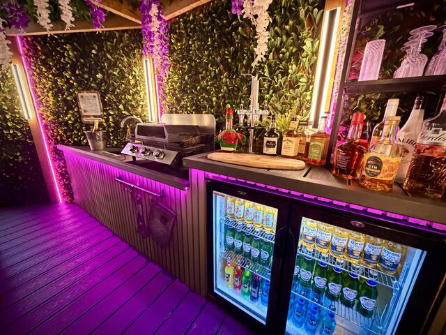 “Stylish pod bar area featuring a fully stocked mini fridge, a countertop prepared with drinks and liqueurs, plus a skillet and hot plate grill ready for outdoor cooking.”