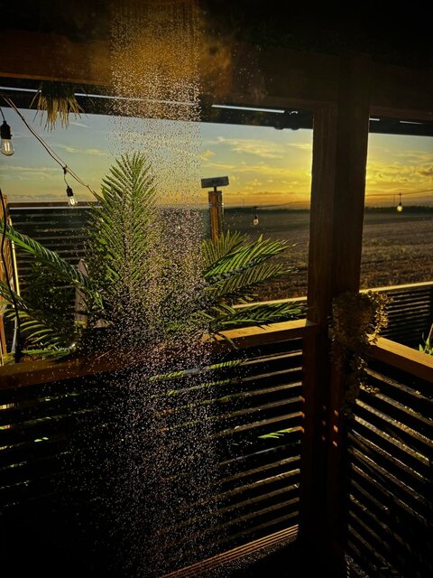 “The Pavilion outdoor shower at The Secret Garden Glamping, with a graceful cascade of water sparkling in the golden sunset. The sky glows deep orange over the surrounding fields.