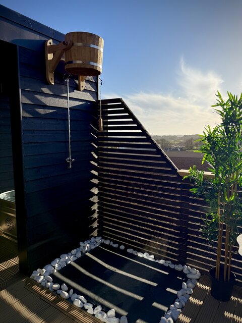 “Outdoor space featuring a luxury Swedish bucket shower, showcased in daylight against sleek jet-black flooring, with crisp white pebbles surrounding the base for a clean, modern glamping aesthetic.”
