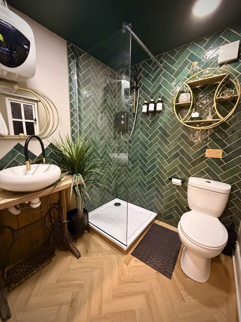 Modern bathroom featuring green tiled walls, beige chevron flooring, a plant beside the shower, an oval sink with a black tap and gold tip, and a stylish mirror at The Secret Garden Glamping.