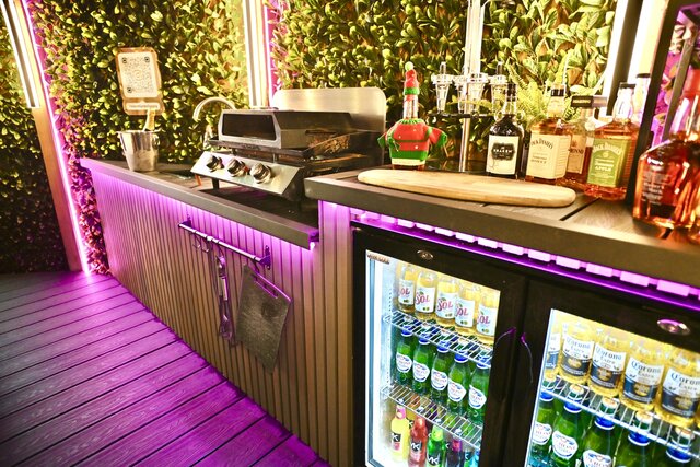 “Stylish pod bar area featuring a fully stocked mini fridge, a countertop prepared with drinks and liqueurs, plus a skillet and hot plate grill ready for outdoor cooking.”