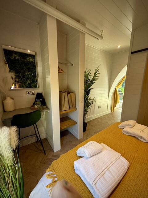 “The Cove bedroom at The Secret Garden Glamping featuring a plush double bed with neatly arranged towels at the foot, set against a lush green plant-covered feature wall, and a stylish dresser.