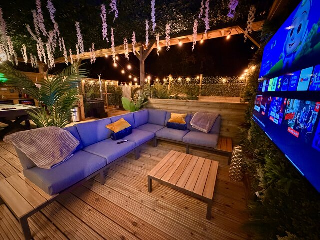 “Nighttime view of the Blossom pod’s outdoor seating at The Secret Garden Glamping, featuring two comfortable couches arranged around a stylish coffee table that matches the golden-brown decking.