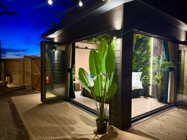 Alternate angle of The Vista bedroom exterior at The Secret Garden Glamping showing the neatly made bed inside, a plant in front, soft white exterior lighting, and a beautiful night sky.