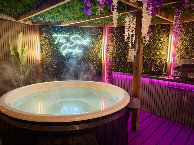 “Luxury hot tub glowing invitingly, designed for total relaxation, with the illuminated Secret Garden Glamping neon sign visible in the background.”