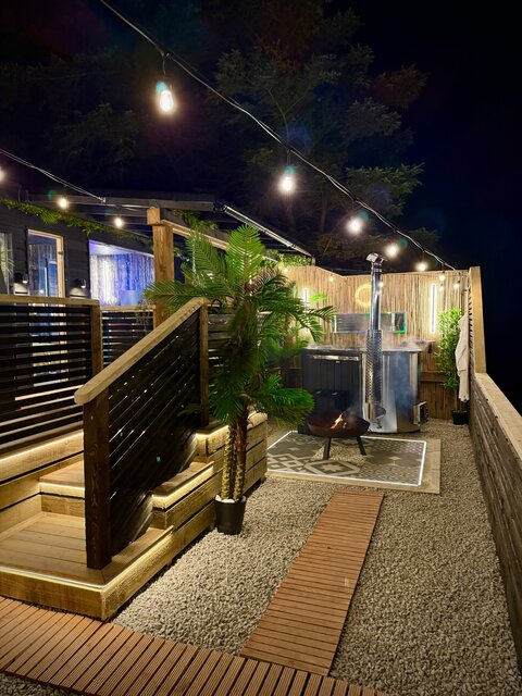 “Exterior night view of the Pavilion unit at The Secret Garden Glamping, featuring the log-burning hot tub in the background and the entire pod beautifully illuminated, creating a warm atmosphere.