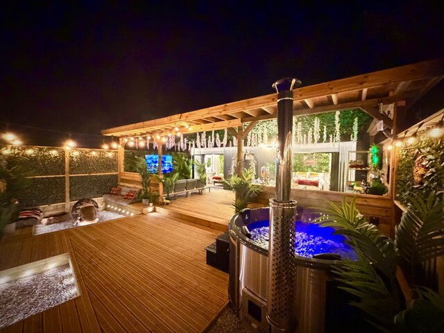 A rustic log-burner hot tub sits on a sweep of golden-brown decking, its water gently steaming into the night air. Just off to the side, a fire pit glows with a lively orange flame.