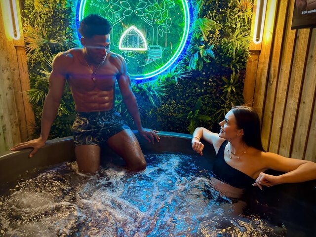 Log burner hot tub set on warm wooden decking inside a private open-fronted hut, featuring a wall-mounted smart TV and a large illuminated Secret Garden Glamping sign, creating a cosy atmosphere.