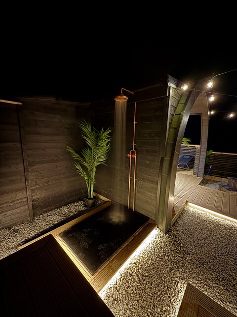 “The Horizon outdoor shower at The Secret Garden Glamping, illuminated with dark neon lighting, creating a moody yet soft ambiance. Water cascades gently, with a lush plant in the corner.