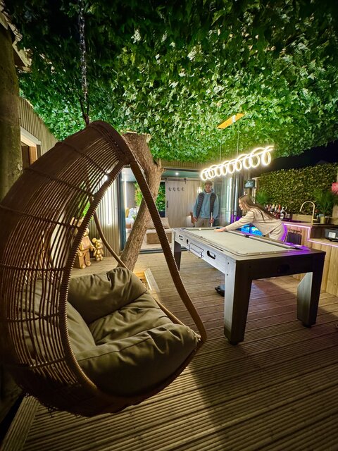 Outdoor oasis with an egg chair, jungle-leaf roof, wooden decking, outdoor kitchen, and a convertible pool/air-hockey/dining table, all accented by swirl lighting and double doors.