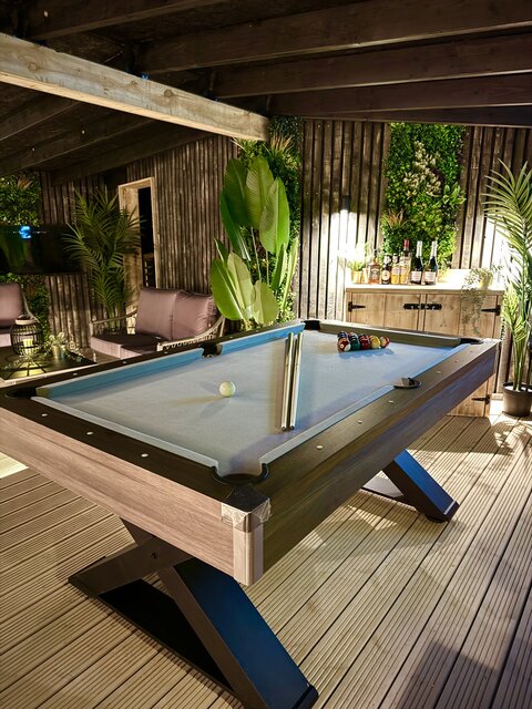 “The Horizon indoor area at The Secret Garden Glamping featuring a well-lit pool table with cues, surrounded by lush greenery. In the background, bottles of champagne ready for celebration.