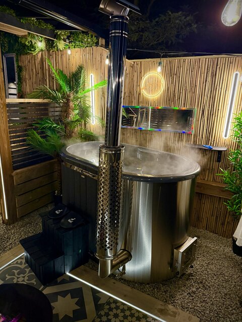 “Night view of the Pavilion at The Secret Garden Glamping featuring a steaming log-burning hot tub, with a plant in the background. The scene radiates warmth, luxury, and a serene atmosphere.
