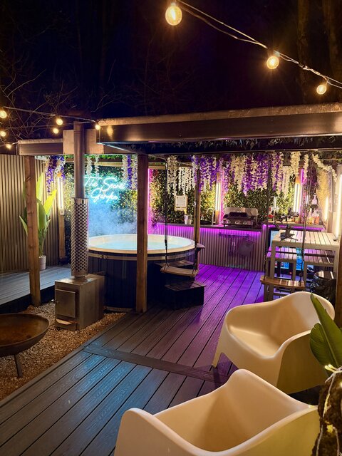 “Luxury outdoor area under the night sky featuring a log-burner hot tub glowing blue, a stylish bar area in the background, and an illuminated Secret Garden Glamping sign creating a warm, upscale atm