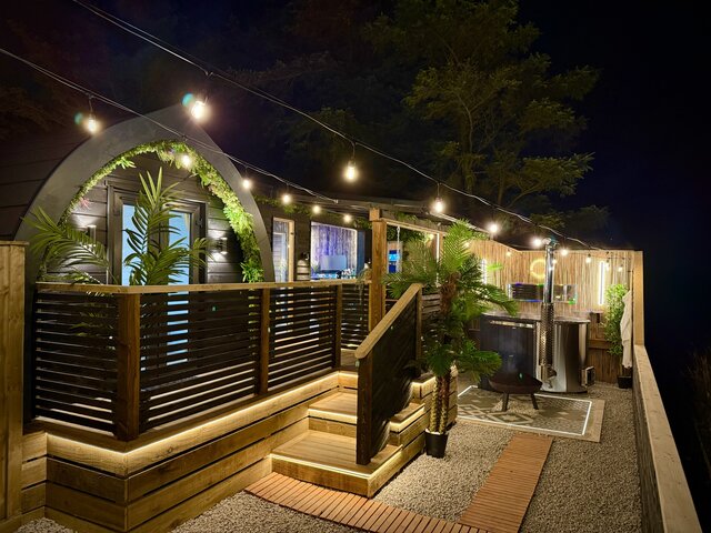 “Outdoor overview of The Pavilion at The Secret Garden Glamping from a unique angle, highlighting the log-burning luxury hot tub tucked into a corner and the pod glowing warmly in the dark night. 