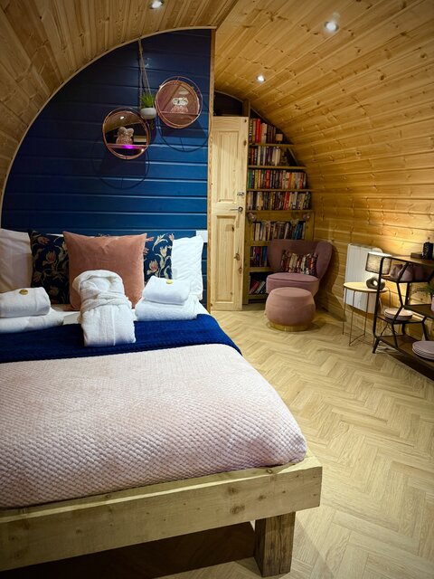 The Glen interior at The Secret Garden Glamping featuring a neatly made double bed with robes and towels, cozy chair with footstool, wooden bookcase, radiator, and two side tables with lamp.