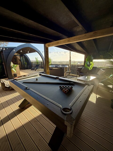 “The Horizon at The Secret Garden Glamping featuring a sleek jet-black pool table set up and ready for play. In the background, the hot tub glows under the warm light of the sunset.