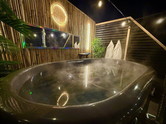 “Nighttime view of a spacious, steaming hot tub at The Secret Garden Glamping, surrounded by soft ambient darkness. Two plush robes hang neatly in the background, 