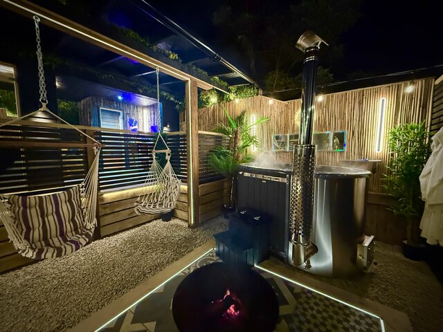 "Close, wide view of The Pavilion outdoor area at The Secret Garden Glamping at night, featuring a steaming log-burning hot tub, bamboo walls and lush plants all creating a luxury day out.