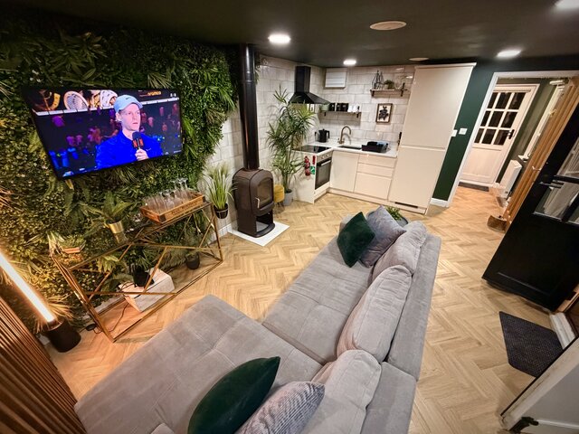 Luxury Lodge living area at The Secret Garden Glamping with beige chevron flooring, half jungle-leaf wall, corner kitchen, smart TV, warm lighting, cozy grey corner sofa, and a log burner fire