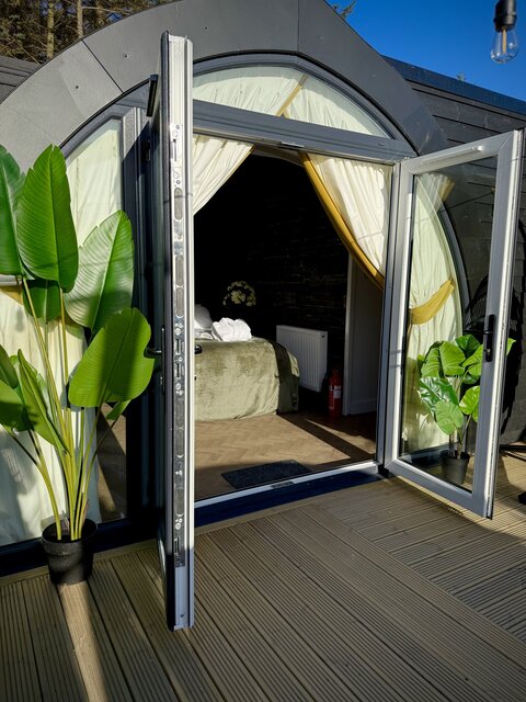 “The Cove bedroom at The Secret Garden Glamping viewed from outside through open doors, featuring an arch-shaped room with a lush green plant on the left.