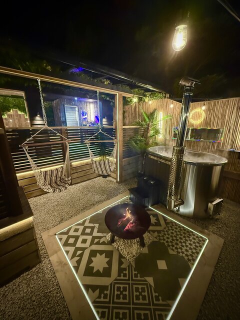 “The Pavilion outdoor area at The Secret Garden Glamping at night, featuring a steaming log-burning hot tub surrounded by bamboo walls and lush plants. 