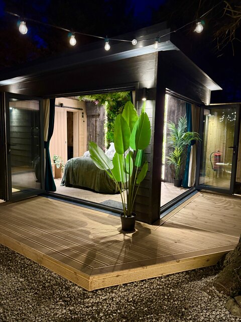 The Vista bedroom at The Secret Garden Glamping viewed from outside, featuring a neatly made bed inside, a plant in front of the interior, and soft white lighting illuminating the exterior.