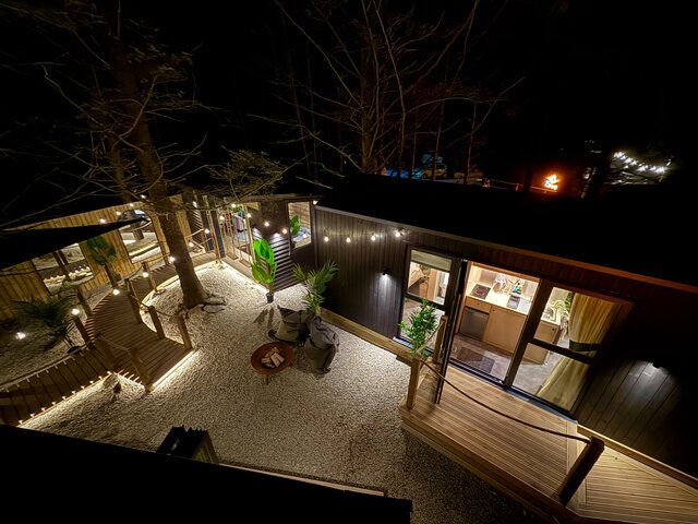 Bird’s-eye view of The Vista outdoor area at The Secret Garden Glamping, showcasing warm string lighting, lush plants, a private hot tub, and a glowing fire feature set on a stony landscaped area.