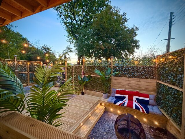 “The Blossom pod outdoor area at The Secret Garden Glamping, featuring green palm-style leaves and carefully placed plants surrounding a firepit ready to be lit. Cushioned seating with cozy blankets 